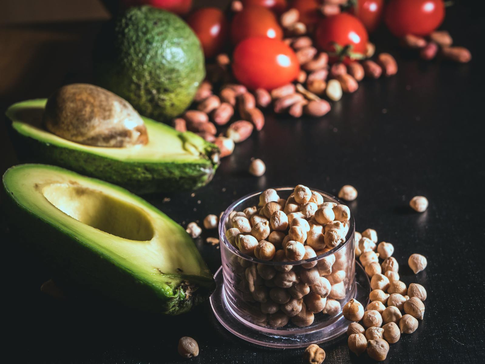 Fresh avocado, chickpeas, nuts, and tomatoes on a dark table, perfect for healthy recipes.