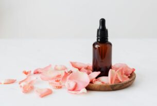 Gentle pale pink wavy rose petals placed on small round wooden plate and table near dark glass essence flask on white background