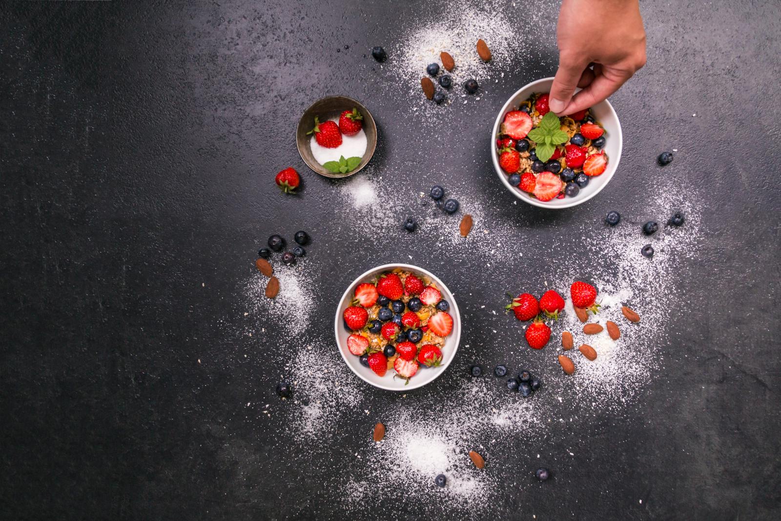 Fresh strawberries, blueberries, almonds in bowls on black table with hand arranging.