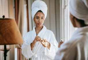 A young woman in a white bathrobe applying skincare cream in front of a mirror.