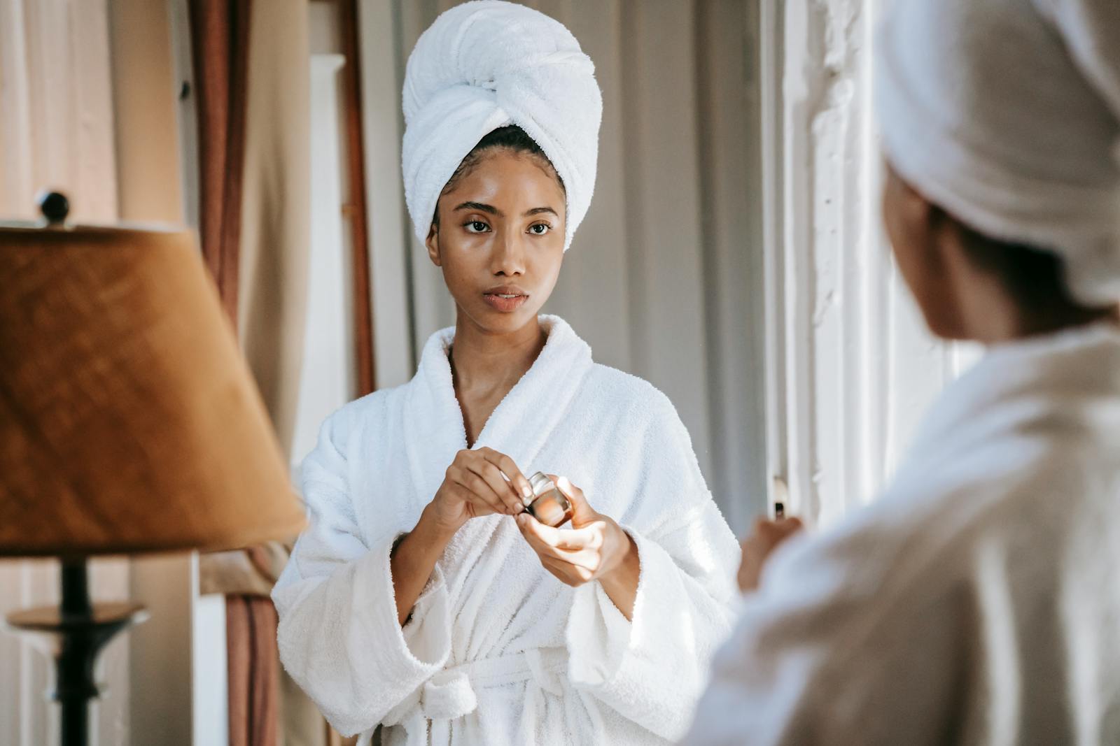 A young woman in a white bathrobe applying skincare cream in front of a mirror.