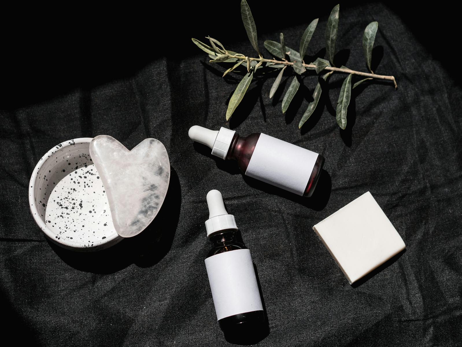 Flat lay of skincare products with gua sha and leaves on black textile. Ideal for beauty and wellness visuals.